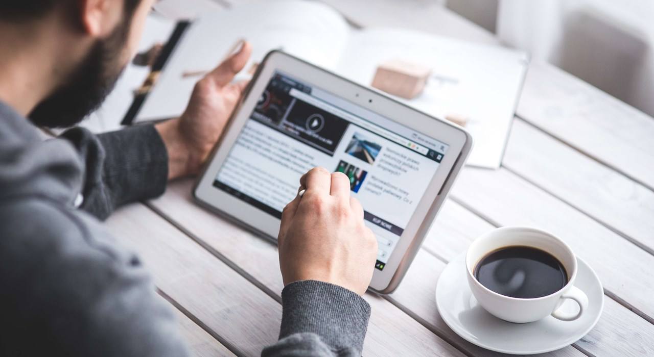 Person using a tablet to browse a website while sitting at a table with a cup of coffee and an open magazine.